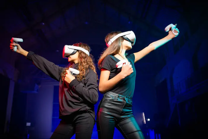 2 girls in VR gear back to back, posing as if they are shooting bows and arrows.